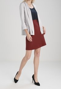 Blazer grigio chiaro con design strutturato, gonna rossa texturizzata e tacchi alti neri. L'outfit presenta un look a strati con colori a contrasto.