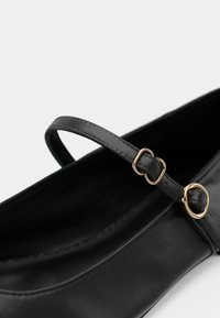 Fekete bőrből készült balerina lapos cipő, vékony pánttal, melyet aranyszínű csat rögzít. Sima textúra és lekerekített orr kialakítás.