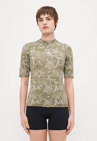 Raglan majica s kratkimi rokavi v olivno zeleni barvi s belim cvetličnim vzorcem, visokim ovratnikom in prilagojenim krojem, v kombinaciji s črnimi kopalkami.