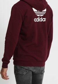 Sudadera con capucha color burdeos con un logo de Adidas blanco y rayas en la parte posterior. Presenta una textura suave y bolsillos laterales.