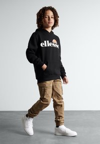 Black hoodie with "ellesse" logo, beige cargo pants with side pockets, and white sneakers. Smooth fabric, relaxed fit, and ribbed cuffs.