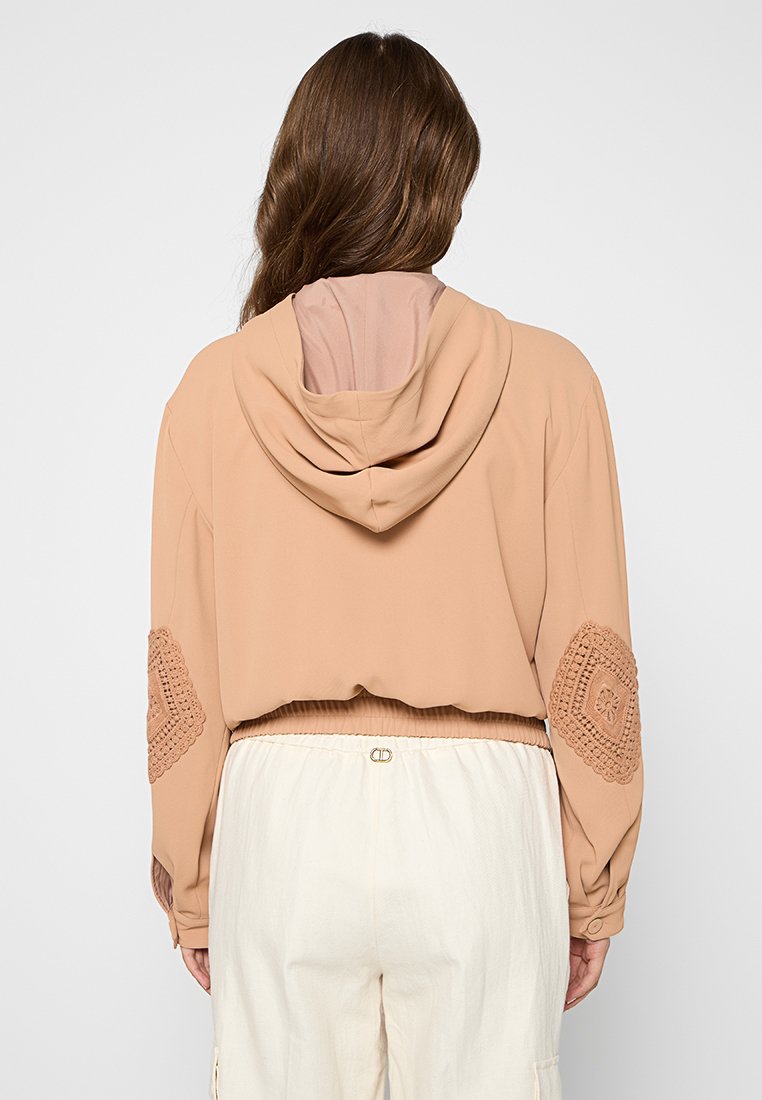 Chaqueta con capucha recortada en beige claro, con acentos de croché texturizados en las mangas y un acabado suave. Presenta un dobladillo ajustado y una tela suave.