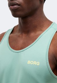 Κοντινό ενός ατόμου που φοράει ανοιχτό πράσινο αμάνικο αθλητικό τοπ με την επιγραφή "BORG" στο στήθος.