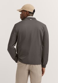 Graue Bomberjacke mit glatter Oberfläche, gerippten Bündchen und Kragen, mit einem dezenten Logodetail auf dem Rücken. Kombiniert mit beigen Hosen.