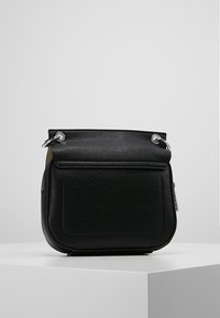 Černá kožená crossbody taška se stříbrnými kovovými kroužky a přední kapsou, umístěná na bílém podstavci v jednoduchém pozadí.