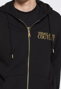 Sudadera con cremallera negra hecha de un tejido suave, con bordado en oro "VERSACE JEANS COUTURE", cordones y un bolsillo frontal.