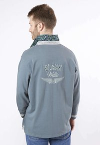 Chemise grise à manches longues avec un col à motifs, arborant les inscriptions "BLACK" et "Wellis" brodées au dos en blanc. Manches à rayures et coupe décontractée.