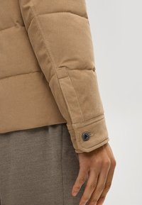 Manica di giacca puffer in corduroy beige con texture trapuntata, dotata di polsino con bottone e design a taglio dritto. Tessuto morbido con cuciture visibili.