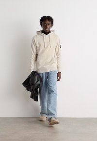 Felpa beige con tasca frontale, jeans in denim azzurro chiaro e scarpe slip-on beige; tiene un giubbotto di pelle nera. Sfondo semplice.