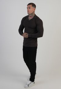 Cămașă polo cu mânecă lungă, de culoare maro închis, cu guler, asortată cu pantaloni negri slim-fit și adidași albi cu accente gri.