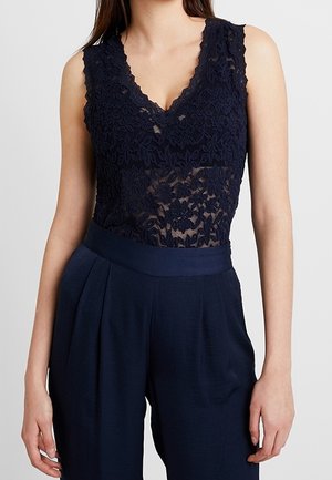 Bodysuit en dentelle bleu marine avec un décolleté en v et des bords festonnés, superposé sur une ceinture en bleu marine unie et un pantalon évasé, présentant des pinces pour ajouter de la texture.