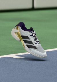 Tennisschuh mit einem weißen Mesh-Obermaterial, lila Akzenten, drei schwarzen Streifen an der Seite und einem goldenen Absatzdetail. Leichtes Design.