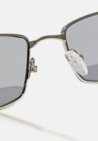Lunettes de soleil avec une monture métallique argentée, verres carrés, verre teinté gris et plaquettes de nez transparentes. Présente un détail de charnière pour l'ajustabilité.