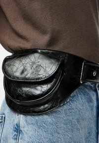 Black, crinkled faux leather fanny pack with a front flap and two compartments, mounted on a denim waistband in a casual outfit.
