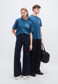 Δύο μοντέλα φορούν μπλε βαμβακερά t-shirts με το λογότυπο "Calvin Klein", σε συνδυασμό με σκούρα τζιν φαρδιάς γραμμής. Το ένα κρατάει μια μαύρη στρογγυλή χειραποσκευή.