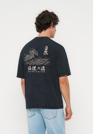 Junger Mann trägt ein schwarzes T-Shirt mit weißem Wellenmotiv und dem Text „WHERE WAVES BECOME LEGENDS“ auf dem Rücken, kombiniert mit hellblauen Jeans.