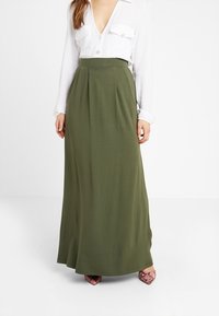 Olive green, floor-length skirt made of soft fabric, featuring a high waist with pleats; worn with a white button-up shirt and pink shoes.