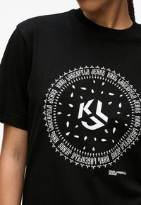 Juodas medvilnės marškinėlis, kuriame yra apskrito balto piešinio su "KL" inicialais ir sudėtingais raštais. Marškinėliai turi trumpas rankoves ir apvalų kaklą.