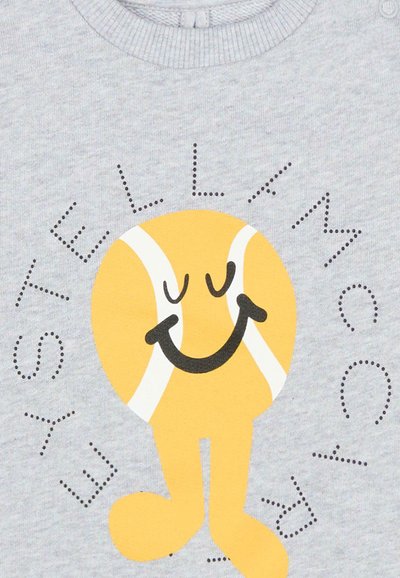 Personaggio di pallina da tennis gialla sorridente con occhi chiusi e gambe su tessuto grigio chiaro, circondato dal testo "ESTELLA MCCARTNEY" in lettere puntinate.