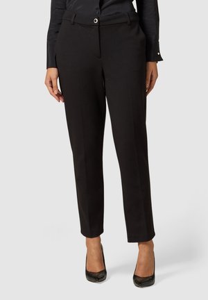 Kobieta w czarnych, dopasowanych spodniach z wysokim stanem i przednimi kieszeniami oraz czarnych szpilkach, na neutralnym tle.