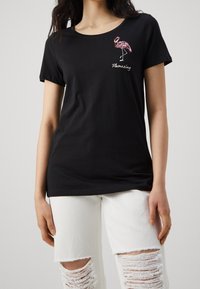 Kvinna i svart t-shirt med en rosa paljetterad flamingo och texten "Flamazing", kombinerat med vita slitna jeans.