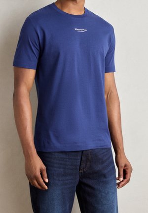 Mann trägt ein schlichtes blaues kurzärmeliges T-Shirt mit kleinem weißen "Marc O'Polo"-Text auf der Brust und blaue Jeans, steht vor einem neutralen Hintergrund.