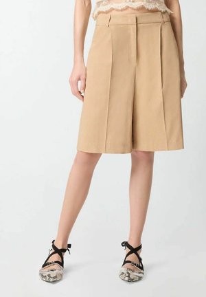 Vrouw draagt beige, op maat gemaakte korte broeken tot aan de knieën en ballet flats met slangenhuidpatroon en zwarte enkelbandjes, staand op een witte achtergrond.