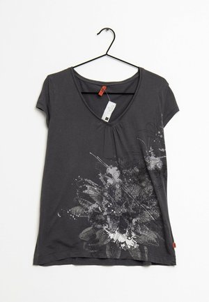 Ciemnoszary T-shirt z krótkim rękawem i dekoltem w serek, z czarno-białym abstrakcyjnym kwiatowym wzorem na dolnej przedniej części, wiszący na czarnym metalowym wieszaku.