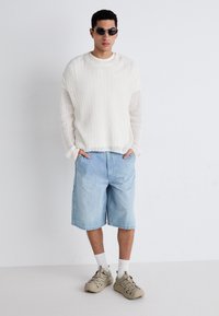 Biała, fakturowana sweter, jasnoniebieskie dżinsowe szorty, białe skarpetki i beżowe sneakersy. Całość dopełniają owalne czarne okulary przeciwsłoneczne.