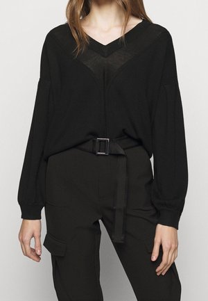 Kvinde iført en sort V-hals sweater med transparente detaljer og sorte bukser med bælte og cargolommer, stående foran en ensfarvet baggrund.