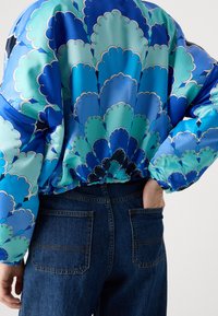 Giacca in satin blu con un motivo scallop in diverse tonalità di blu e verde, aderente in vita, abbinata a jeans in denim scuro.