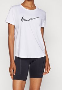 Camiseta atlética de manga corta blanca con el logotipo negro de Nike, combinada con shorts de ciclismo negros que presentan sutiles detalles de cremallera. Textura de tela suave.