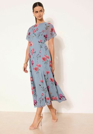 Frau steht vor einem hellen Hintergrund, trägt ein blaues Midikleid mit pinkem und rotem Blumenmuster sowie hautfarbene High-Heel-Sandalen.