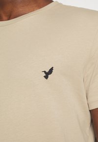 Beige T-shirt made of soft fabric, featuring a round neckline and short sleeves. Contains small black hummingbird embroidery on the chest.