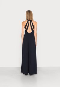 Marinblå halterneck jumpsuit med öppen rygg, utrustad med flera remmar och en flödande, bredbenad design. Tillverkad av ett mjukt, strukturerat tyg.