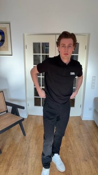 Junger Mann steht drinnen mit den Händen in der Hüfte, trägt ein schwarzes Karl Lagerfeld Polohemd, schwarze Hose und weiße Turnschuhe.