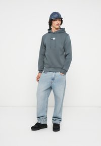 Felpa grigia con coulisse regolabile, logo blu a contrasto, jeans larghi azzurri, e scarpe nere; indossata con un cappello blu.