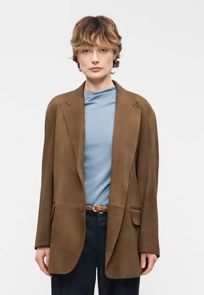 Personne aux cheveux courts et ondulés portant des boucles d'oreilles créoles dorées, un haut bleu clair, un blazer marron surdimensionné, une ceinture marron et un pantalon foncé, debout devant un fond blanc.