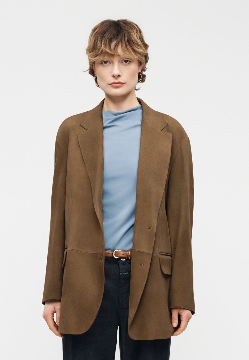 Personne aux cheveux courts et ondulés portant des boucles d'oreilles créoles dorées, un haut bleu clair, un blazer marron surdimensionné, une ceinture marron et un pantalon foncé, debout devant un fond blanc.