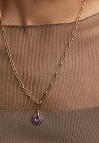 Collar de cadena de eslabones en tono dorado con un colgante circular morado, que presenta un agujero central. En el fondo, una blusa texturizada color beige.