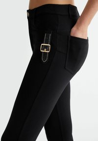 Melnas bikses, kas izgatavotas no elastīga auduma, ar detalizētu sānu kabatu, kas dekorēta ar sprādzi un rotājumiem. Formas piegrieziens un gluda tekstūra.