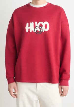 Person iført en rød sweatshirt med lange ærmer med hvid "HUGO" tekst og et sort smiley-ansigt på brystet, parret med lyse bukser.