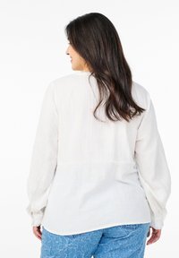 Långärmad, vit blus i ett lätt material med en strukturerad yta. Har rund halsringning och en sömdetalj vid midjan.