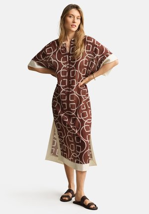 Vrouw draagt een bruine midi-jurk met een wit koordpatroon, witte mouwboorden en zoom, staat met handen in de zij, draagt zwarte sandalen.