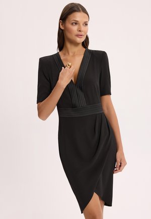 Kobieta ubrana w czarną sukienkę do kolan z krótkimi rękawami, głębokim dekoltem w serek, plisowanymi detalami i asymetrycznym dołem, trzymająca złoty pierścionek.