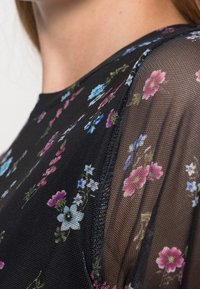 Schwarzes Oberteil aus floralem Netz mit kurzen Ärmeln, das mit kleinen pinken, blauen und weißen Blumen verziert ist, sowie einem runden Ausschnitt mit strukturiertem Rand.