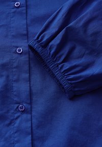 Blusa blu con apertura frontale a bottoni, polsini elasticizzati e texture del tessuto liscia. Presenta bottoni abbinati e colletto da camicia.