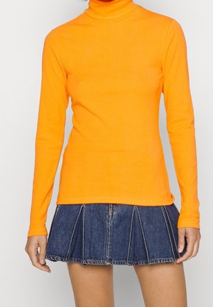 Žena v jasně oranžovém roláku s dlouhými rukávy a tmavě modré plisované džínové minisukni stojí před jednobarevným pozadím.