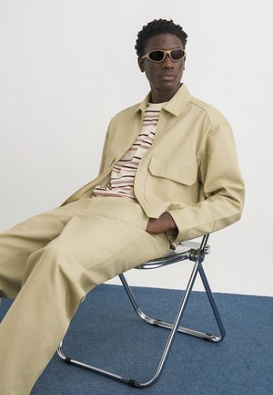 Mężczyzna ubrany w beżową kurtkę i spodnie, w paskowaną koszulę i okulary przeciwsłoneczne, siedzi na składanym krześle z metalowym stelażem na niebieskim dywanie.