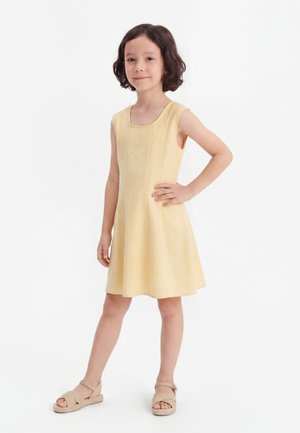 Νεαρό κορίτσι στέκεται με αυτοπεποίθηση φορώντας ένα κίτρινο φόρεμα χωρίς μανίκια και μπεζ σανδάλια σε λευκό φόντο.